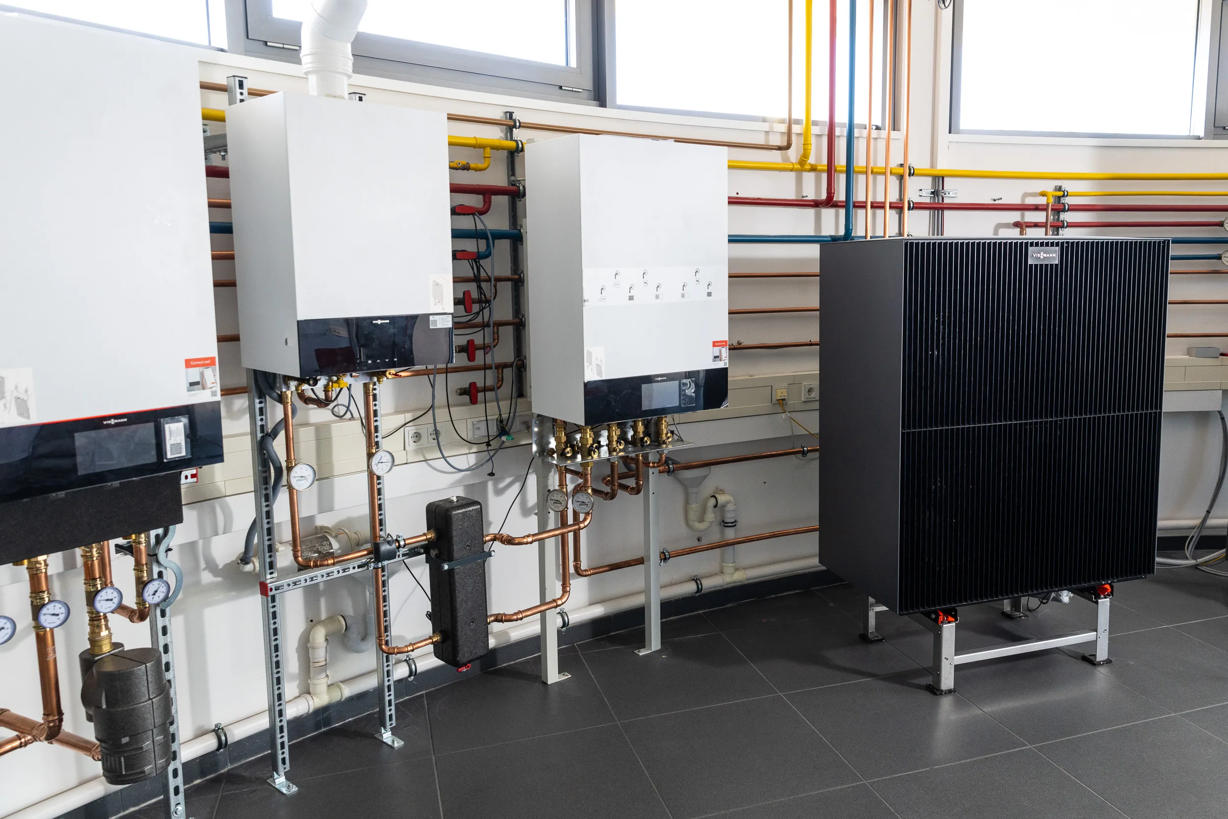 Werkstattwand mit drei modernen Viessmann-Wandgeräten und einer Wärmepumpen-Außeneinheit, angeschlossen an ein umfangreiches Kupfer- und Kunststoff-Rohrsystem.