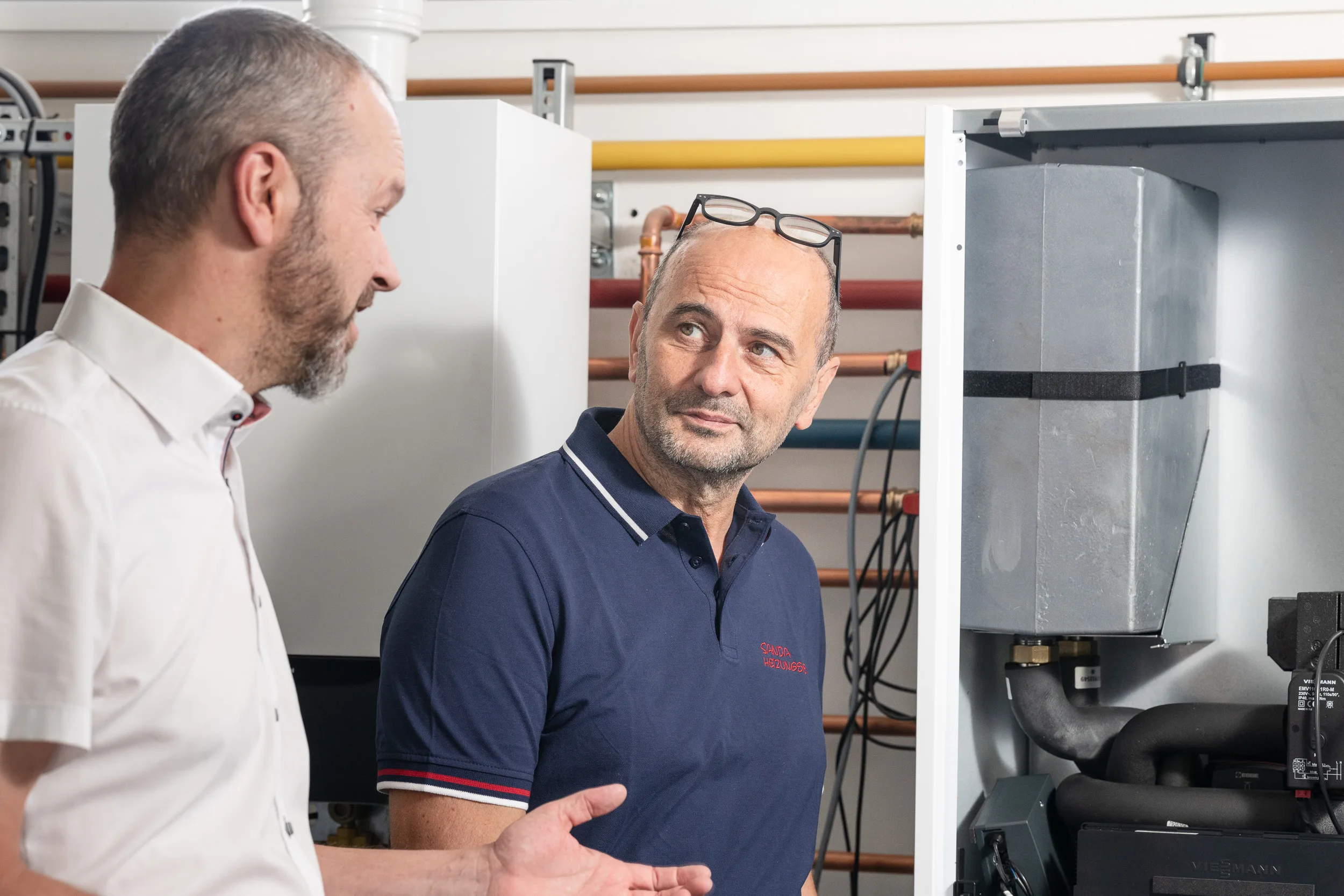 Fachgespräch neben einer geöffneten Viessmann-Heizungsanlage mit Kupferverrohrung, einer der Gesprächspartner trägt ein dunkelblaues Sanda-Heizungsbau-Poloshirt.